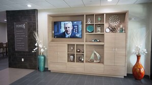 A modern living room with a flat-screen TV, wooden cabinets, decorative items, and a sign for 'HOME2'. - Home2 Suites by Hilton Tulsa Hills (Tulsa)