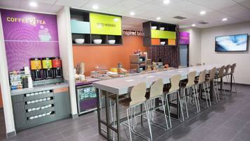 Una estación moderna de café y té con una barra alargada, varios dispensadores de bebidas y un área de descanso con sillas y mesas.