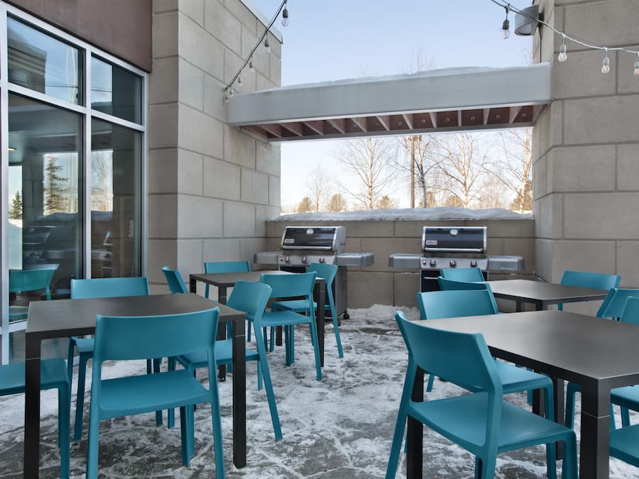 An outdoor seating area with tables and chairs, a barbecue grill, and a view of trees through a window.