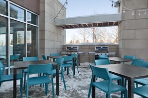 An outdoor seating area with tables and chairs, a barbecue grill, and a view of trees through a window. - Home2 Suites by Hilton Anchorage / Midtown (Anchorage)