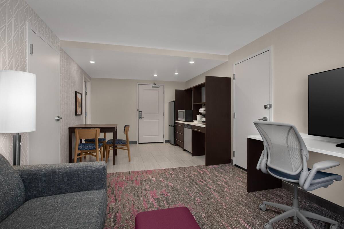 A hotel room with a sofa, a dining table with chairs, a kitchenette, a TV, and a desk with a chair.