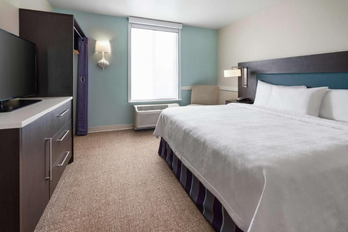 A hotel room with a bed, a TV, a desk, a chair, a window, and a wall-mounted lamp.