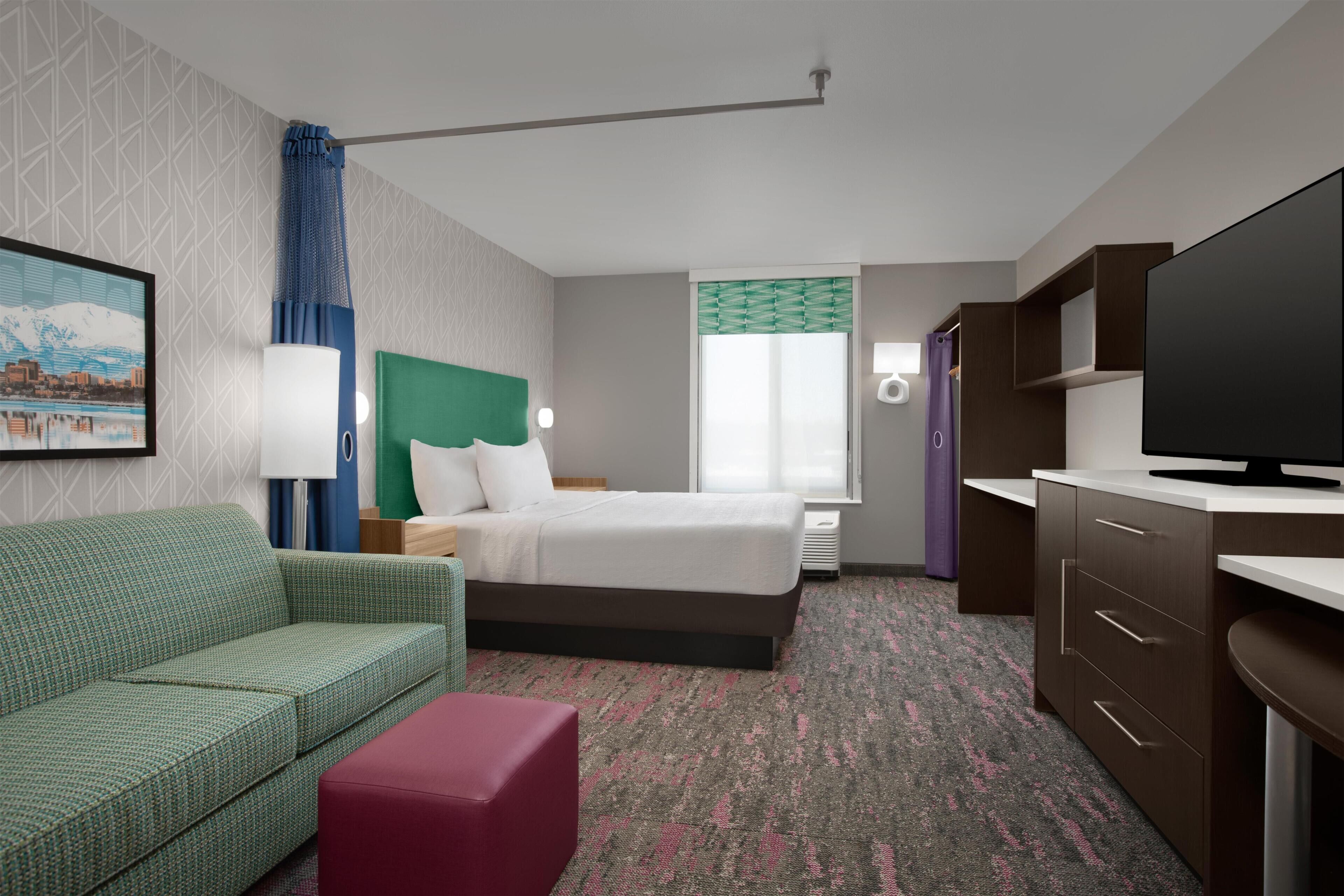 A hotel room with a bed, a green sofa, a red ottoman, a television, a desk, and a window with curtains.