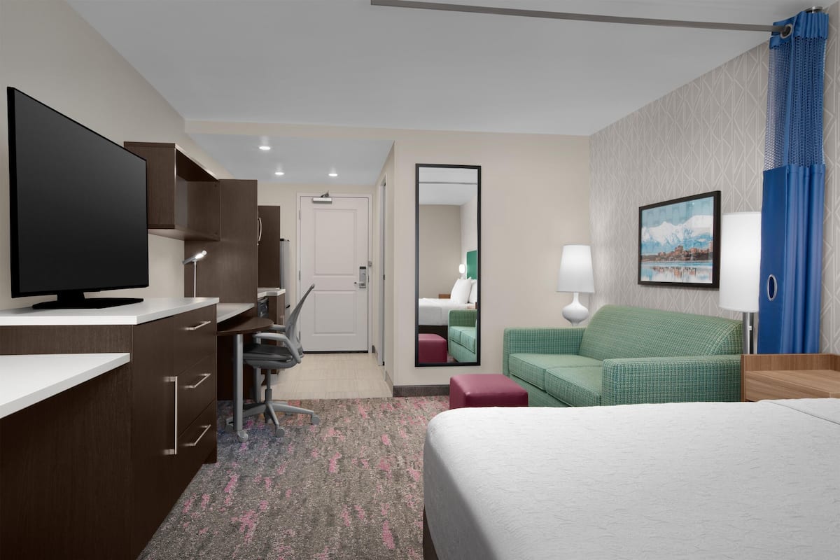 A hotel room with a bed, a desk with a TV, a green sofa, a red ottoman, and a bathroom visible through an open door.