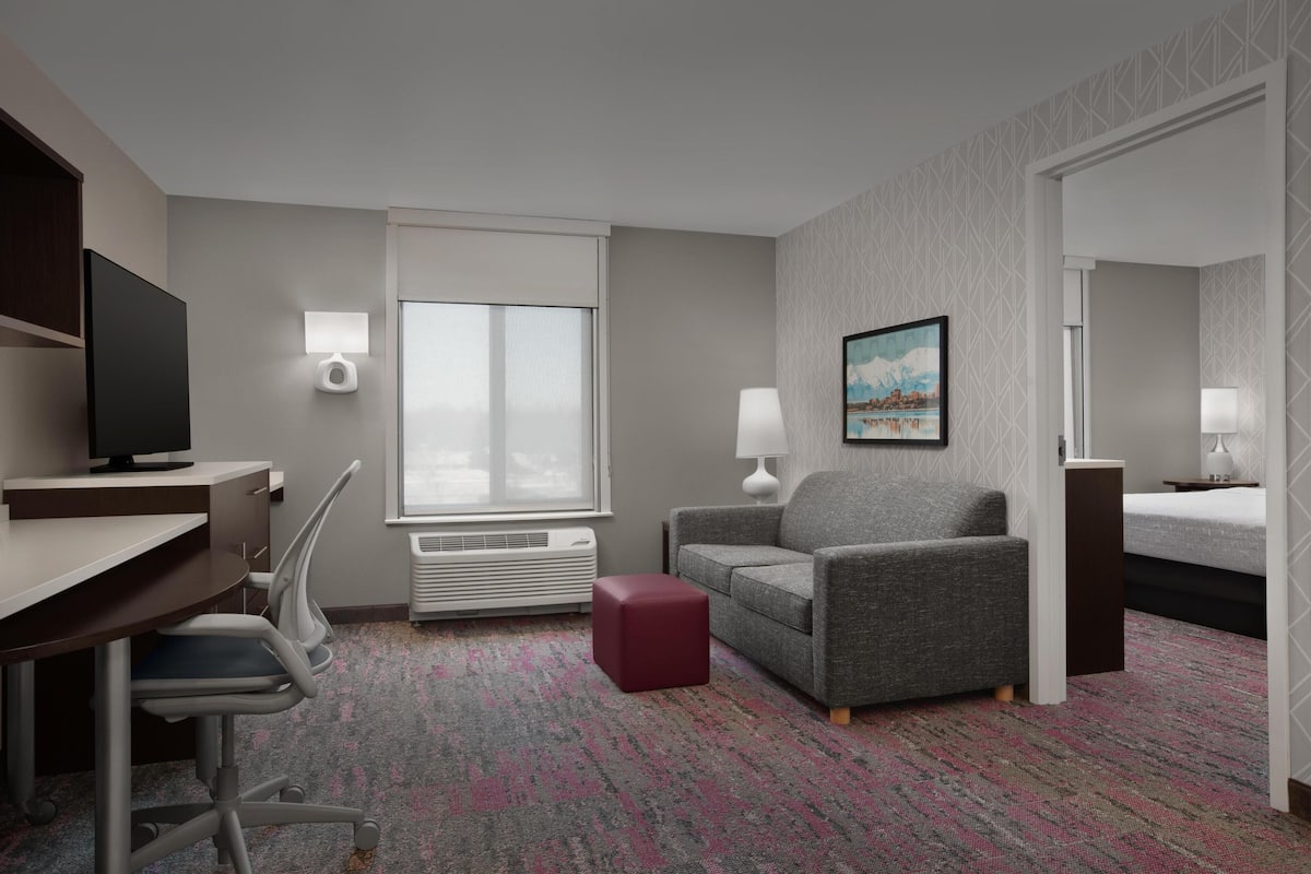 A hotel room with a desk, chair, grey sofa, red ottoman, and a bed.