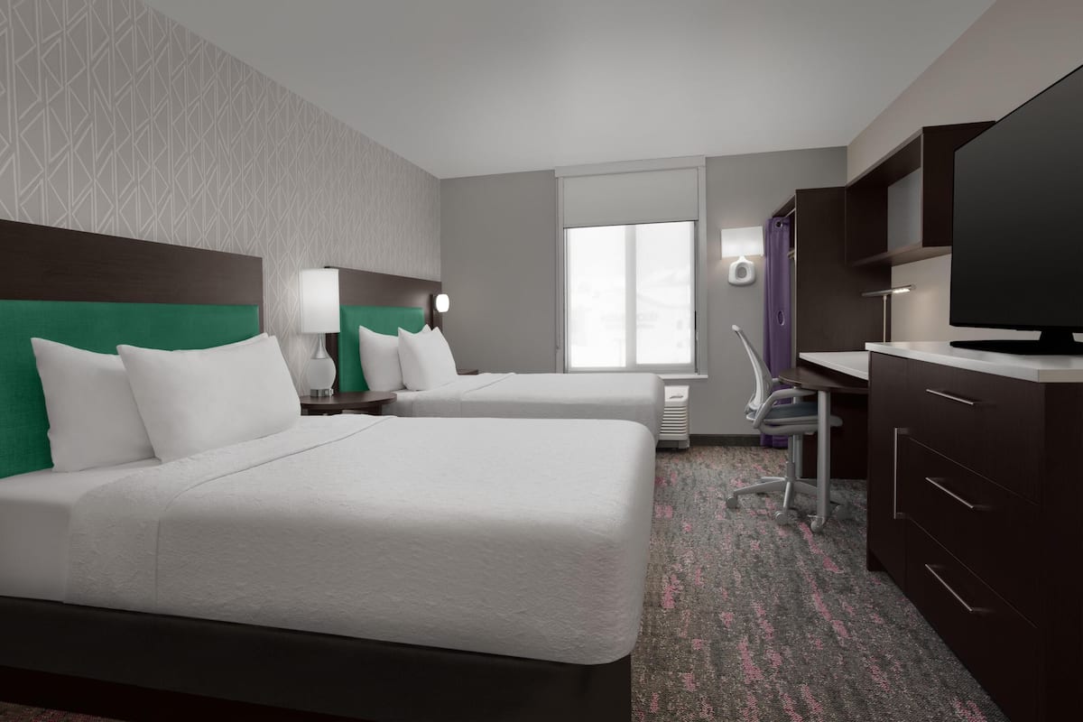 A hotel room with two beds, a desk, a television, and a window.