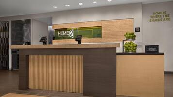 Taulell de recepció amb un cartell que diu 'HOME 2', un cartell de 'Hilton Honors' i una paret amb la frase 'HOME IS WHERE THE SNACKS ARE'.