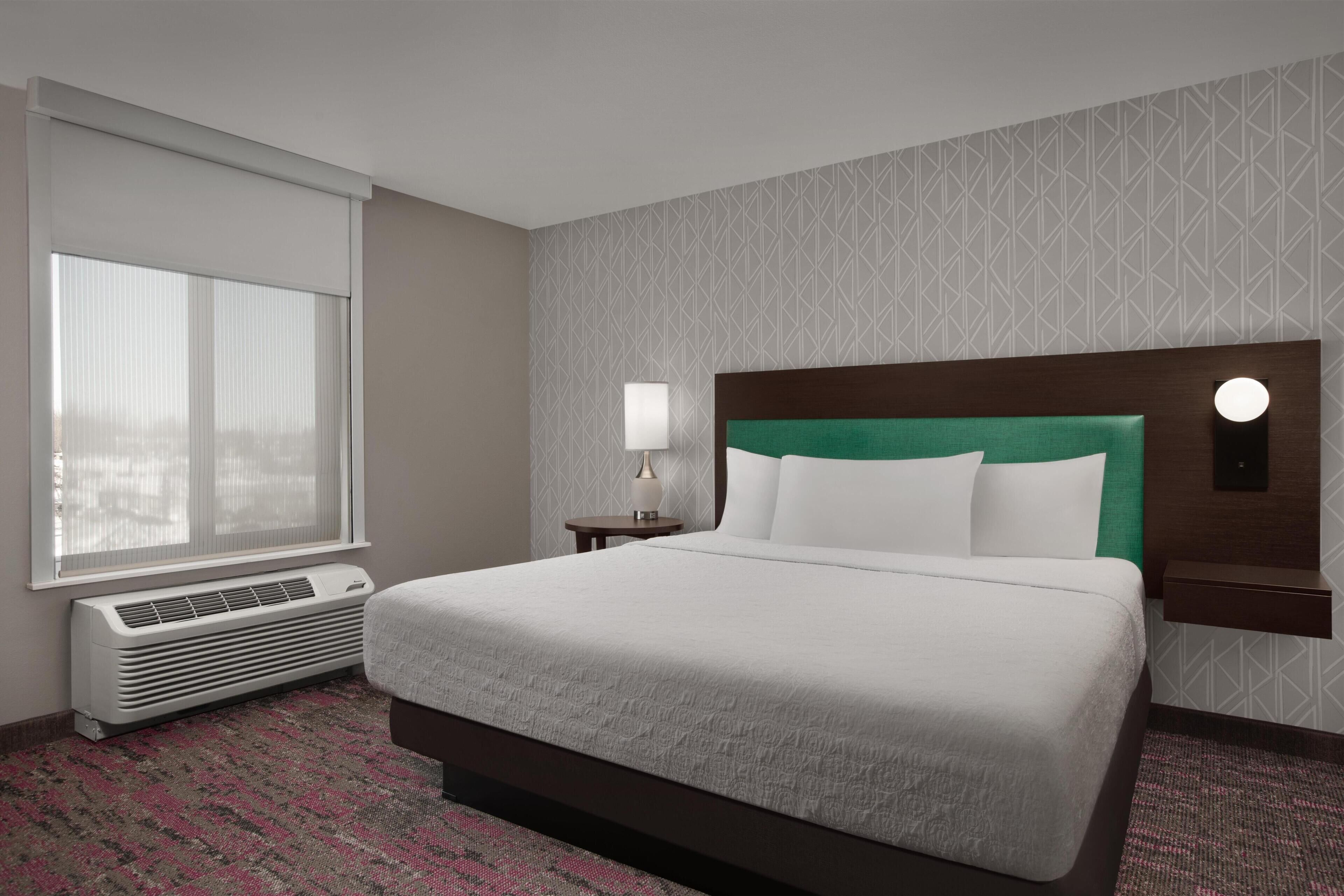 A hotel room with a bed, a window with blinds, a wall-mounted heater, a nightstand with a lamp, and a patterned carpet.