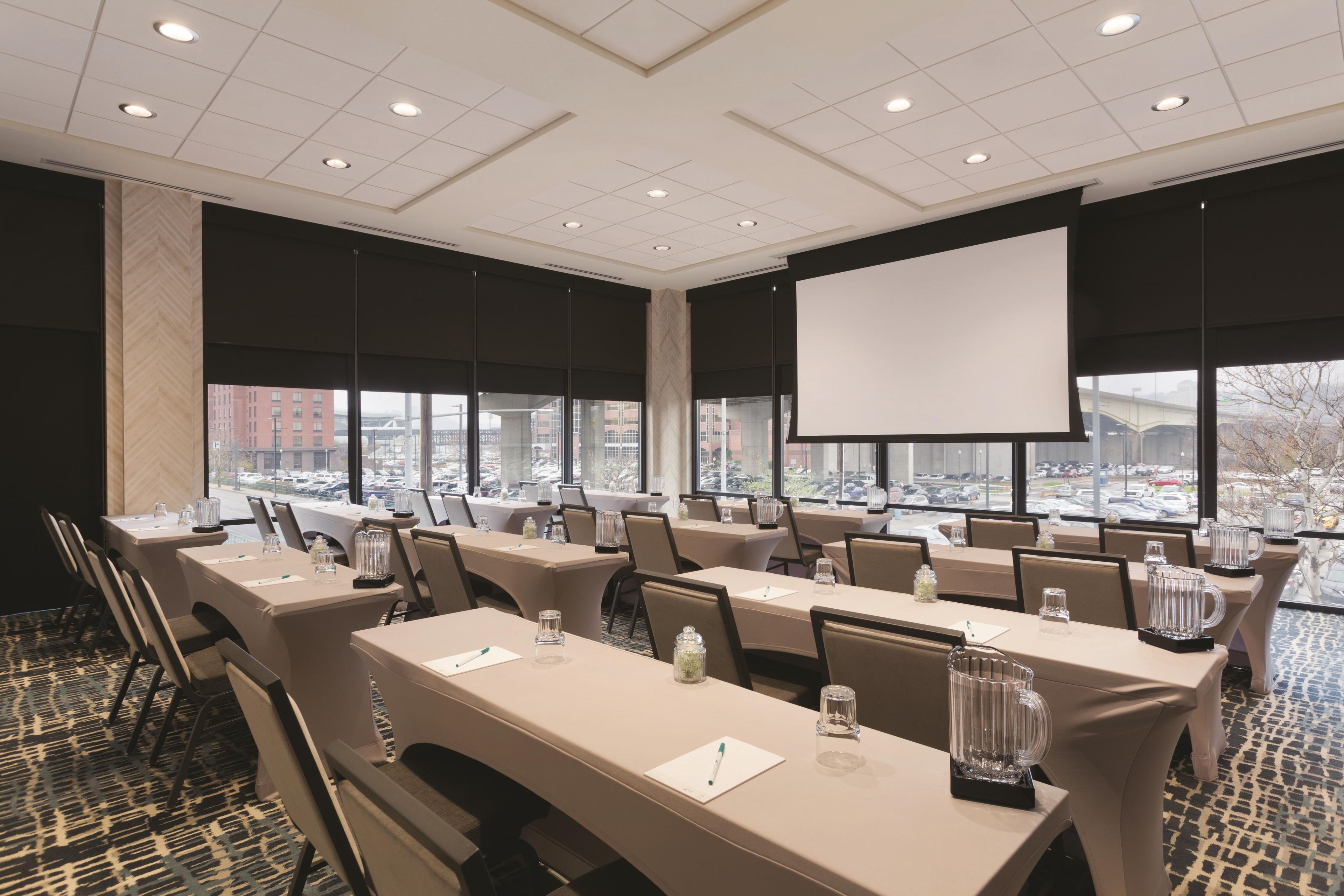 A conference room with multiple tables, chairs, a projector screen, and large windows with a view of a parking lot.