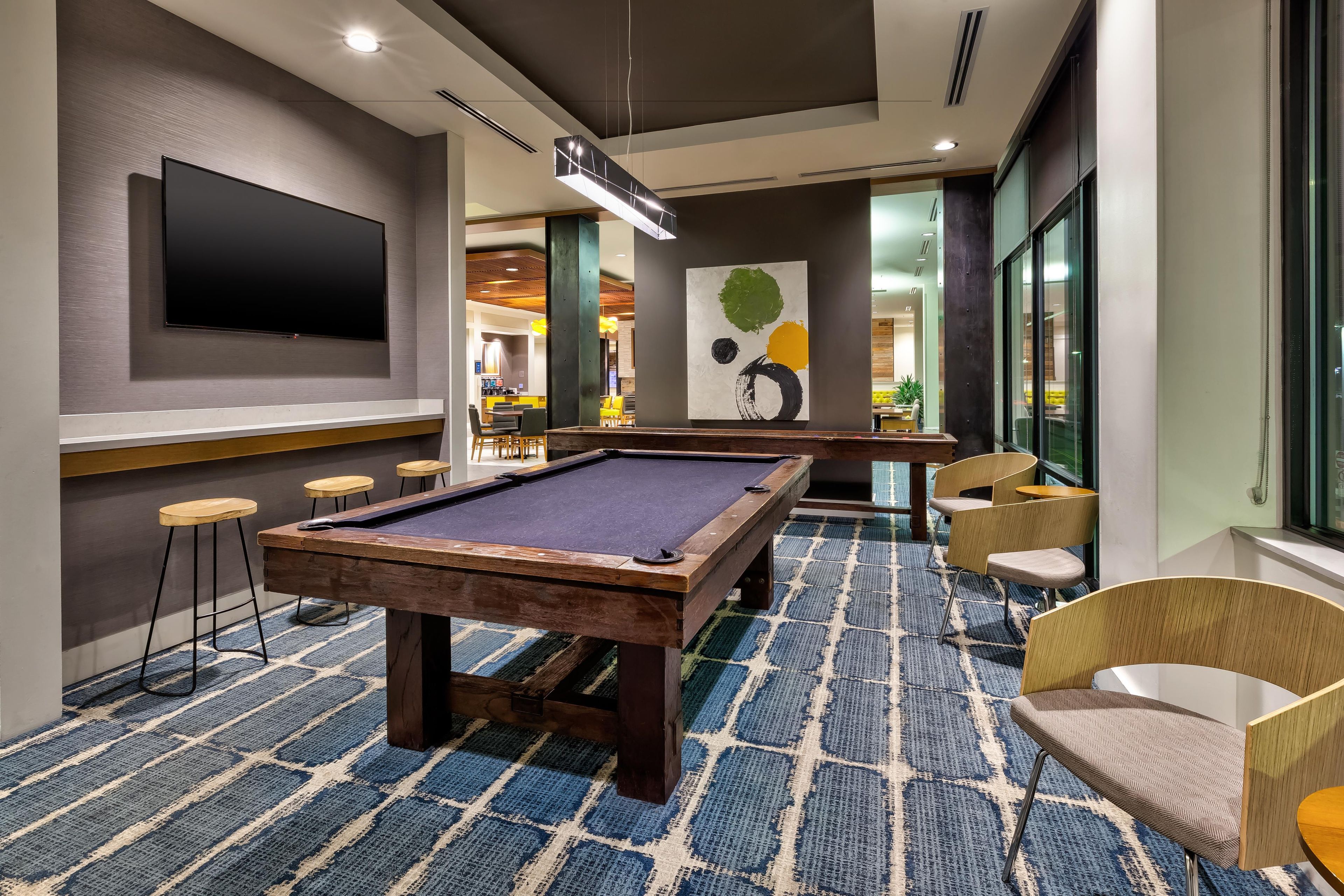 A pool table in a room with a patterned carpet, a flat-screen TV on the wall, and a painting.