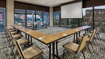 Sala de conferências com uma mesa comprida, cadeiras, tela de projetor e janelas amplas com vista para a cidade.