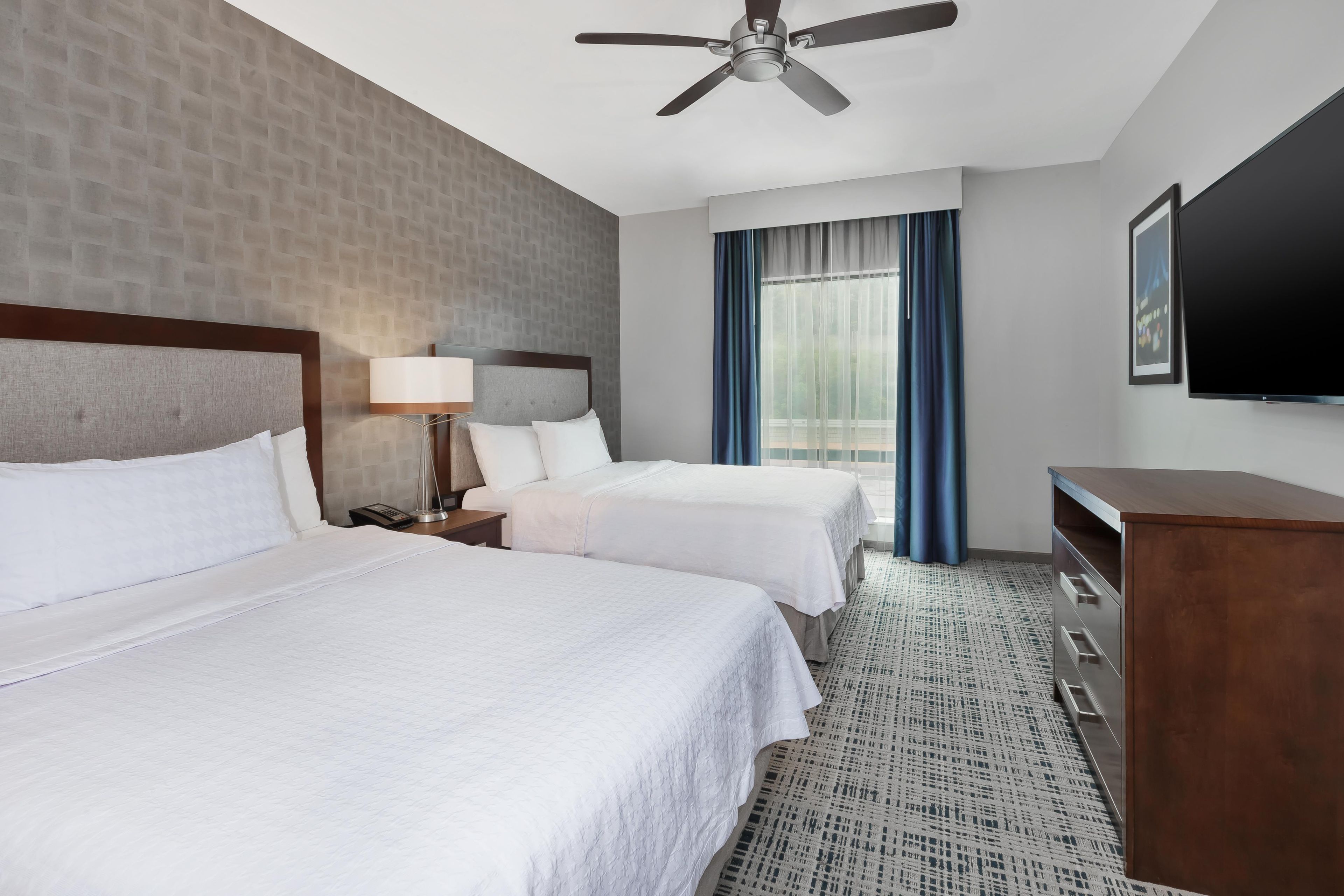 Une chambre d’hôtel avec deux lits, un ventilateur de plafond, un bureau avec une lampe, un téléviseur à écran plat et une grande fenêtre avec des rideaux.