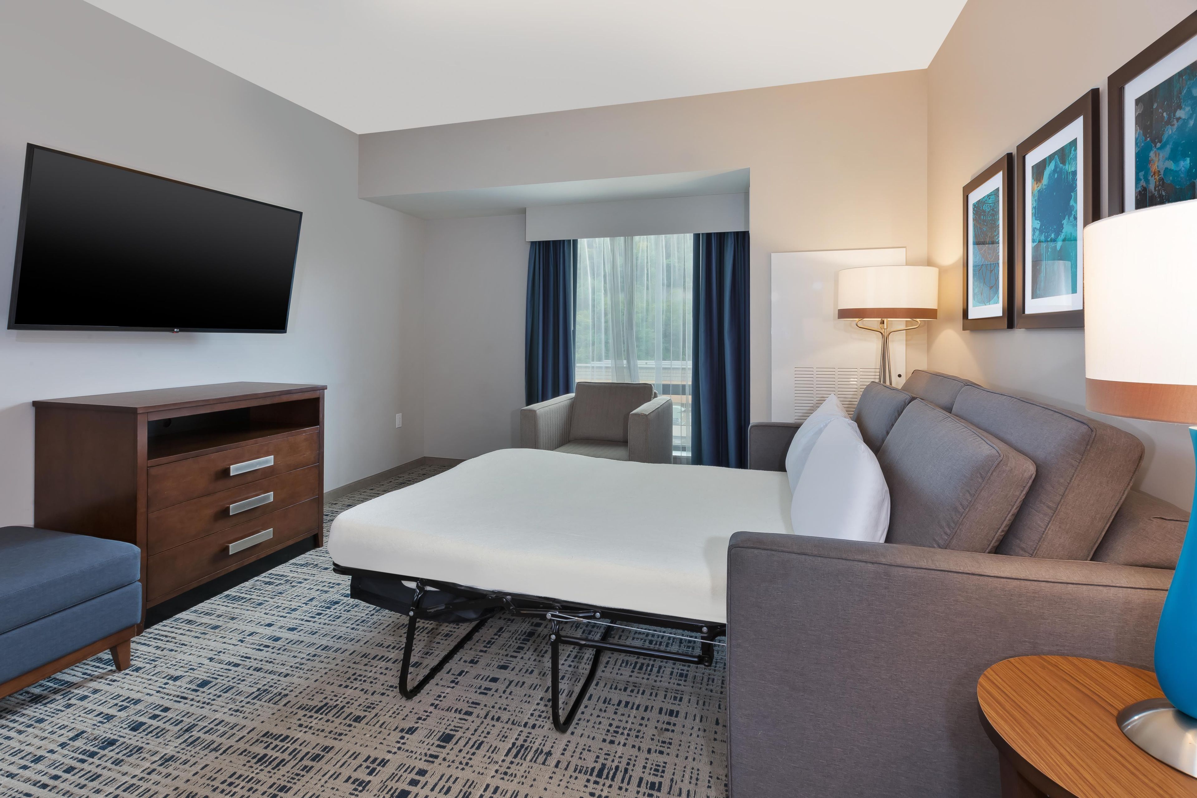 Une chambre d’hôtel avec un lit, un bureau, une télévision, une lampe et une fenêtre avec des rideaux.