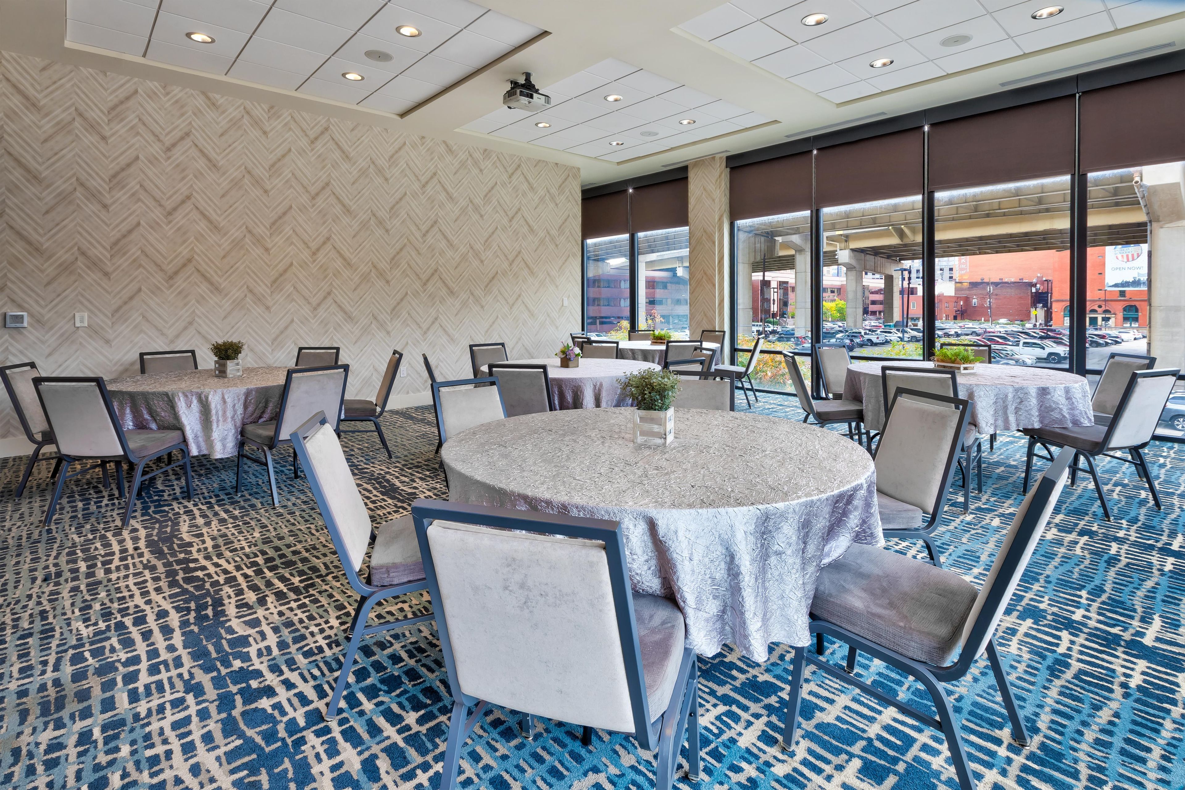 A conference room with multiple tables and chairs, large windows with a view of the city, and a patterned carpet.
