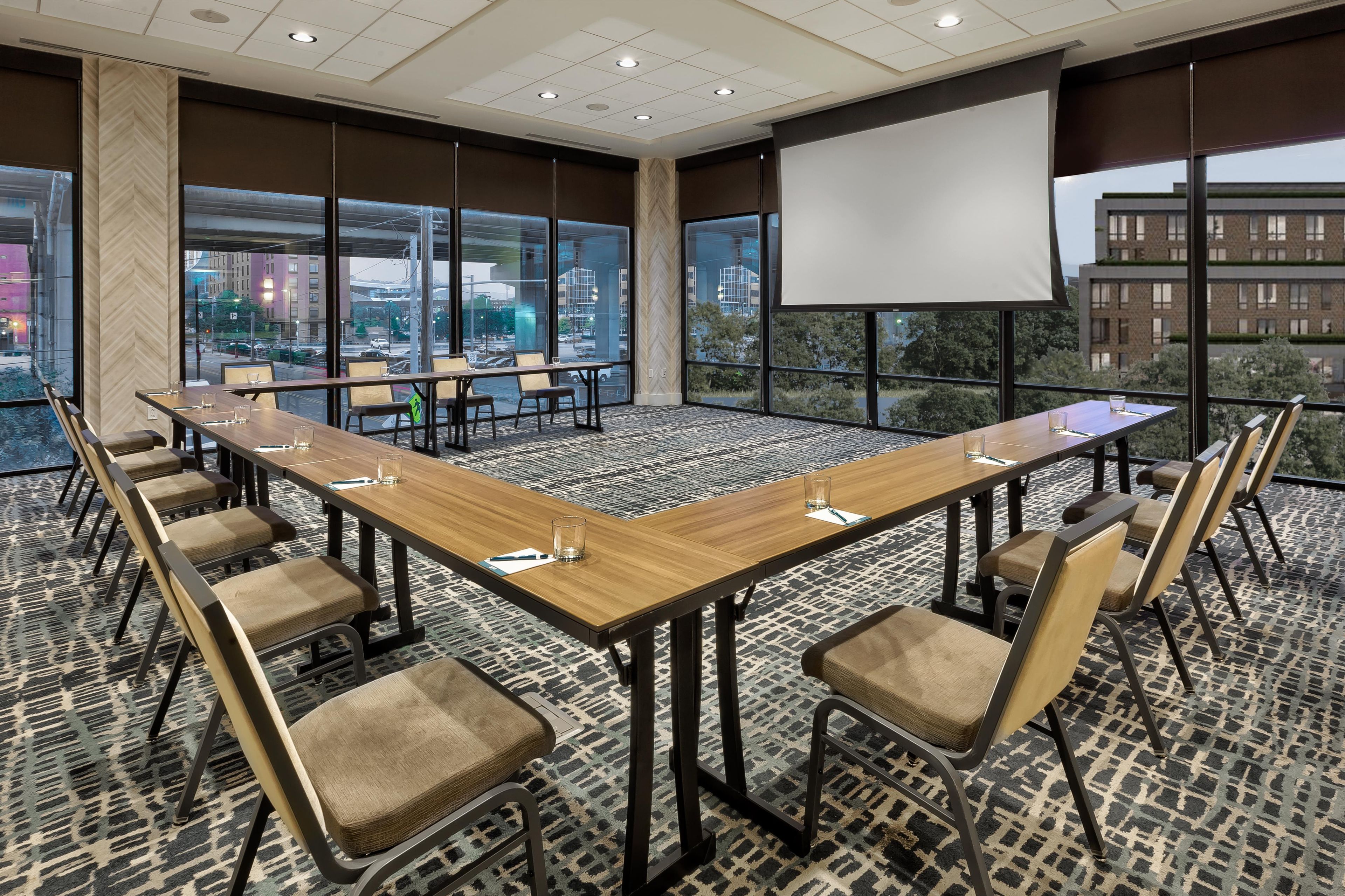 Un salón de conferencias con una larga mesa, sillas, una pantalla de proyector y ventanas grandes con vistas a la ciudad.