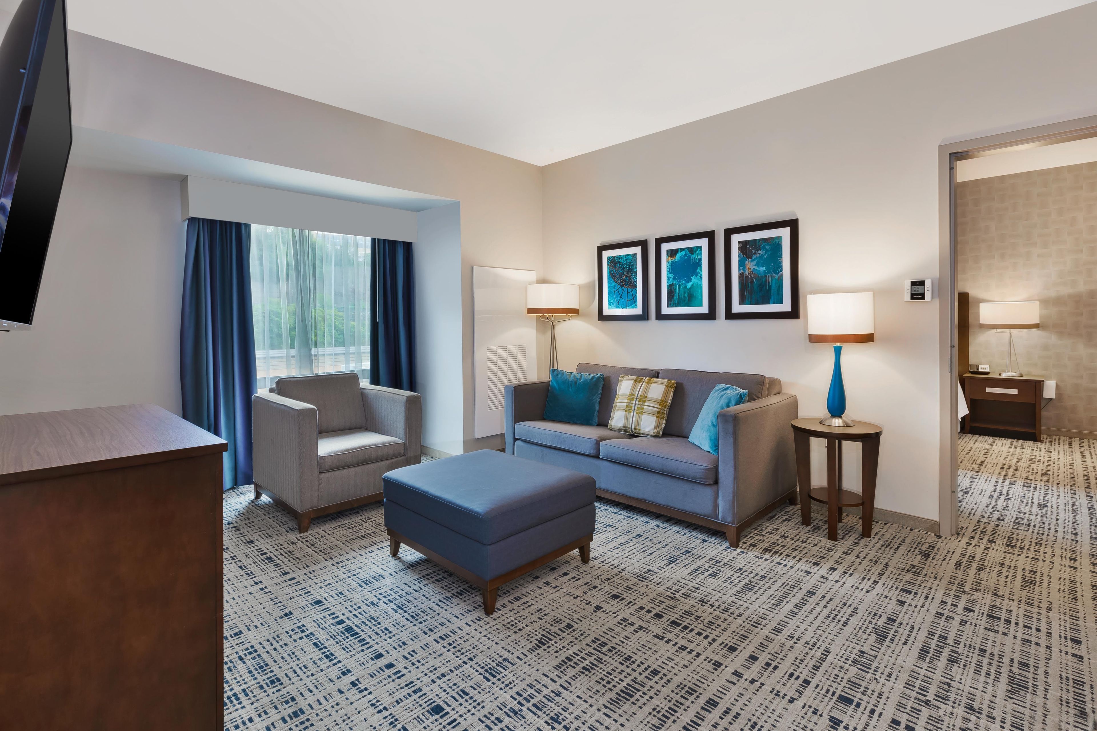Habitación de hotel con sofá, sillón, reposapiés y televisor de pantalla plana.