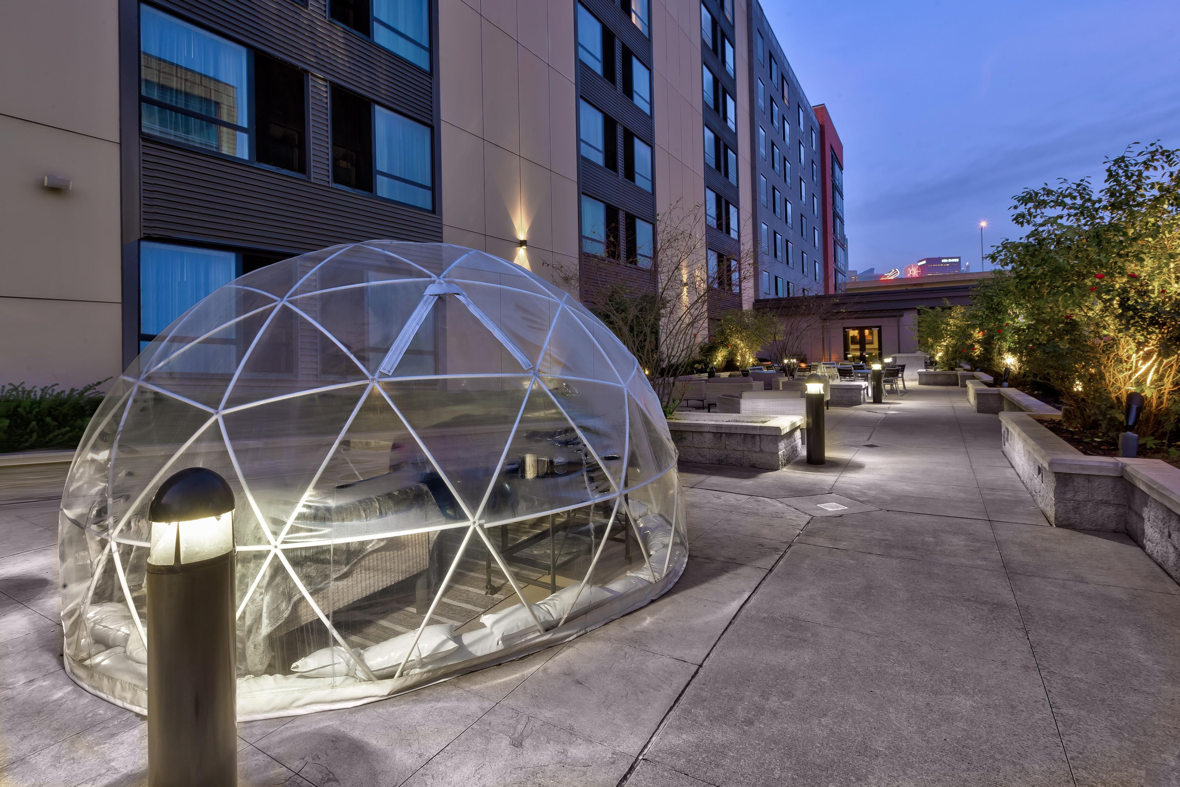 Une structure en forme de dôme géodésique transparente, avec des sièges à l’intérieur, située sur une promenade pavée, entourée de bâtiments et éclairée par des lampadaires.