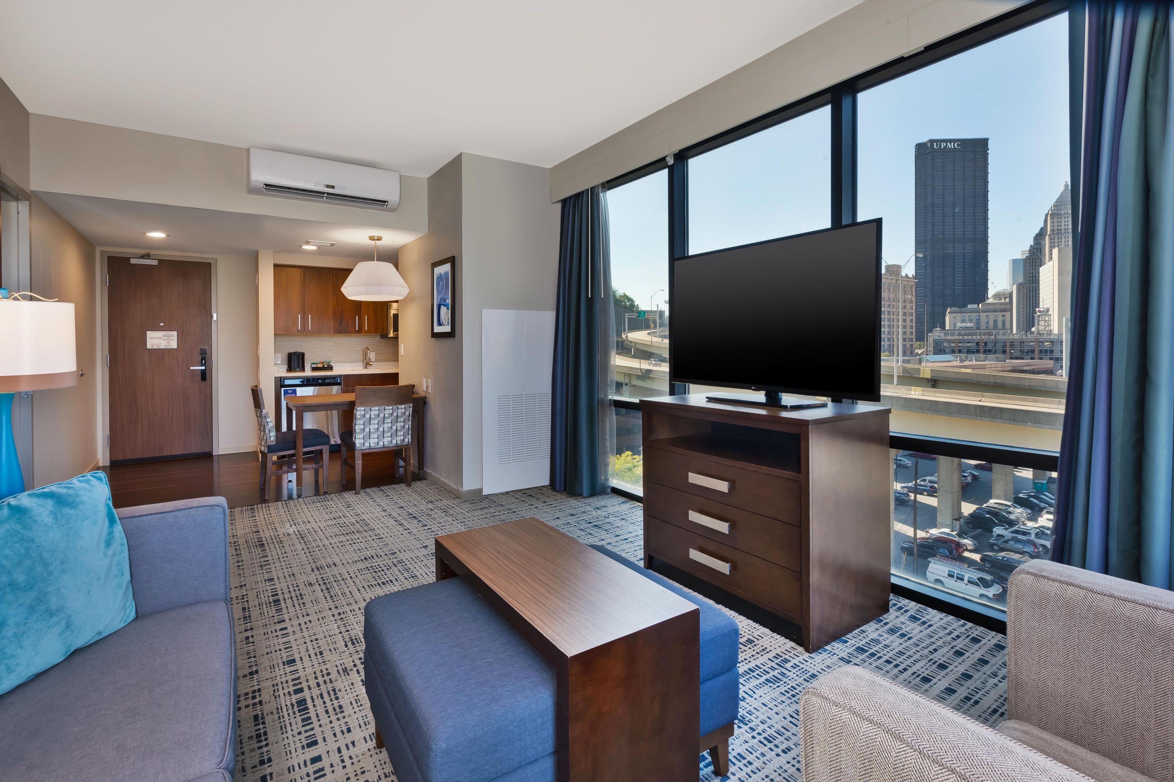 Hotelkamer met een groot raam, een flatscreen televisie, een bank, een salontafel, een eethoek met een keuken op de achtergrond, en uitzicht over de stad.
