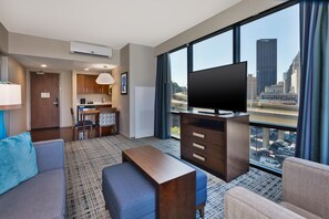 A hotel room with a large window, a flat-screen TV, a sofa, a coffee table, a dining area with a kitchen in the background, and a city view.