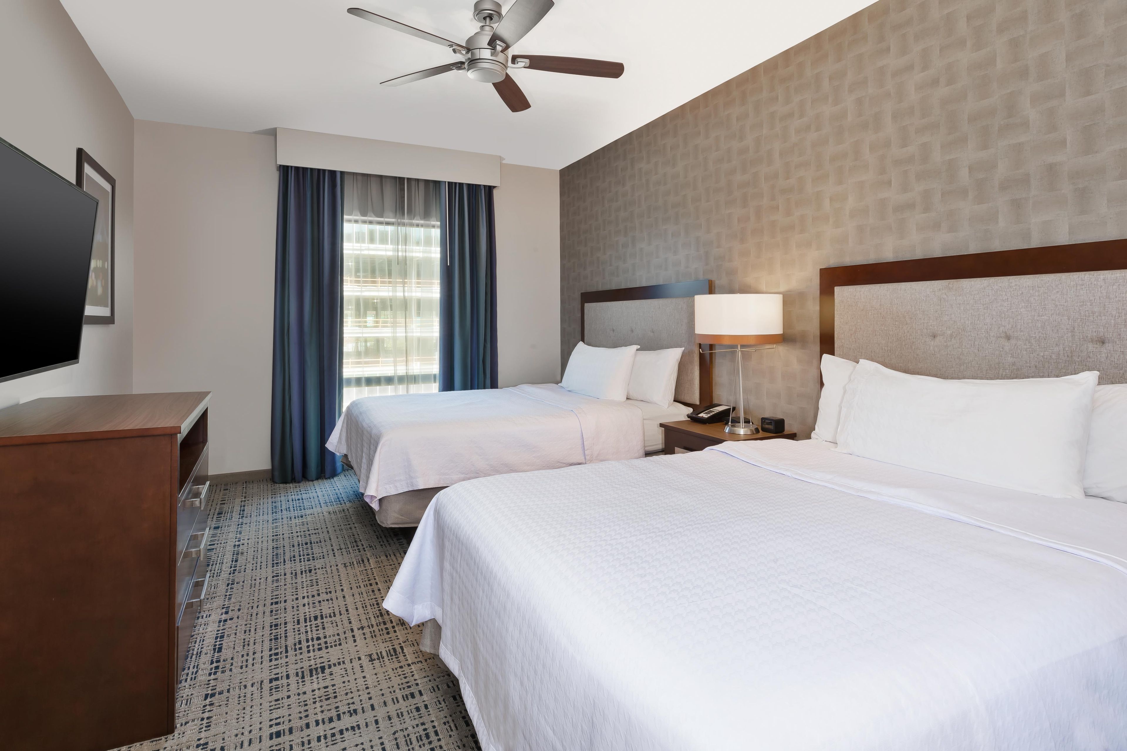 A hotel room with two beds, a ceiling fan, a TV, and a window with curtains.