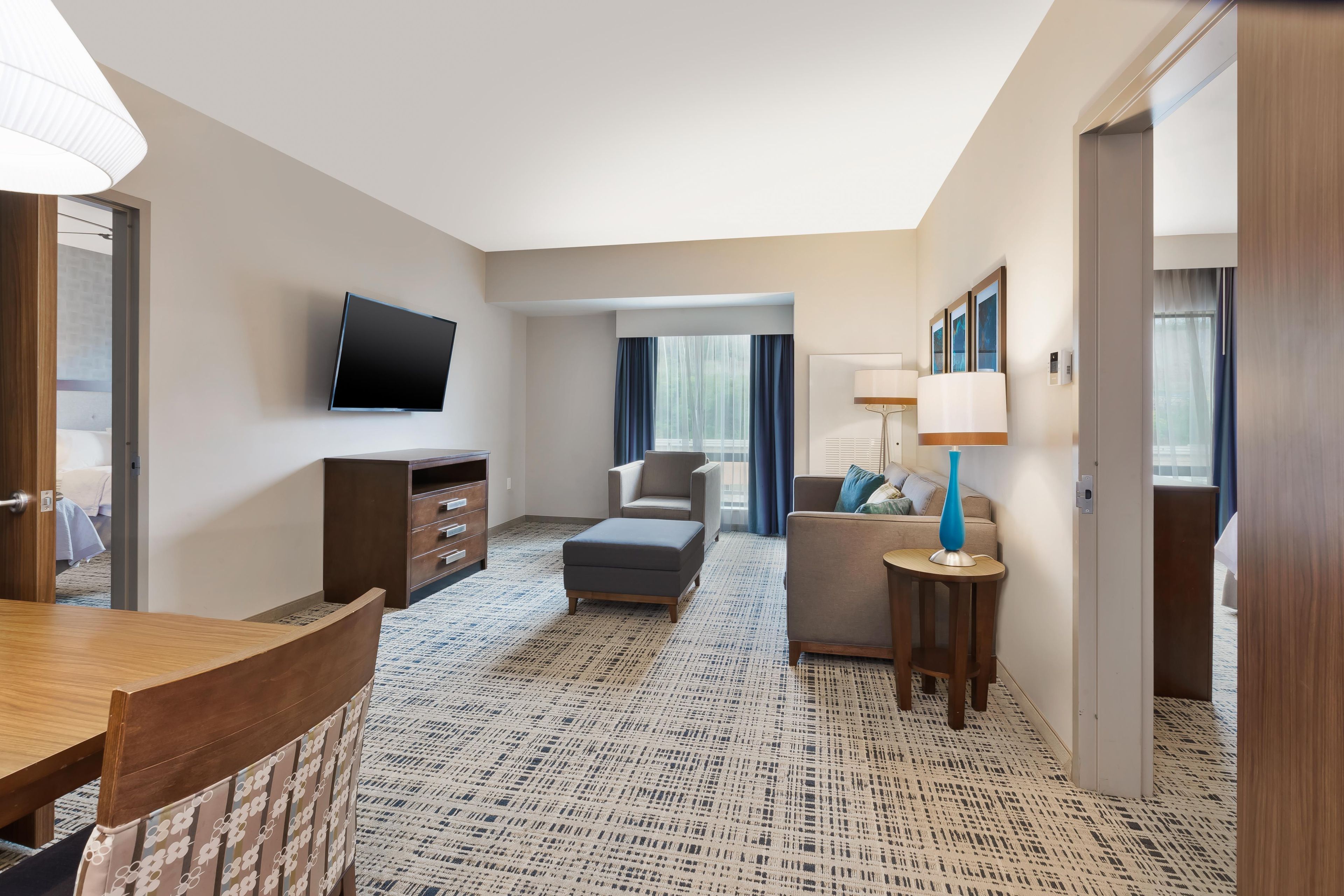 Une chambre d’hôtel dotée d’un téléviseur à écran plat fixé au mur, d’un canapé, d’une petite table avec une lampe et d’un tapis à motifs.
