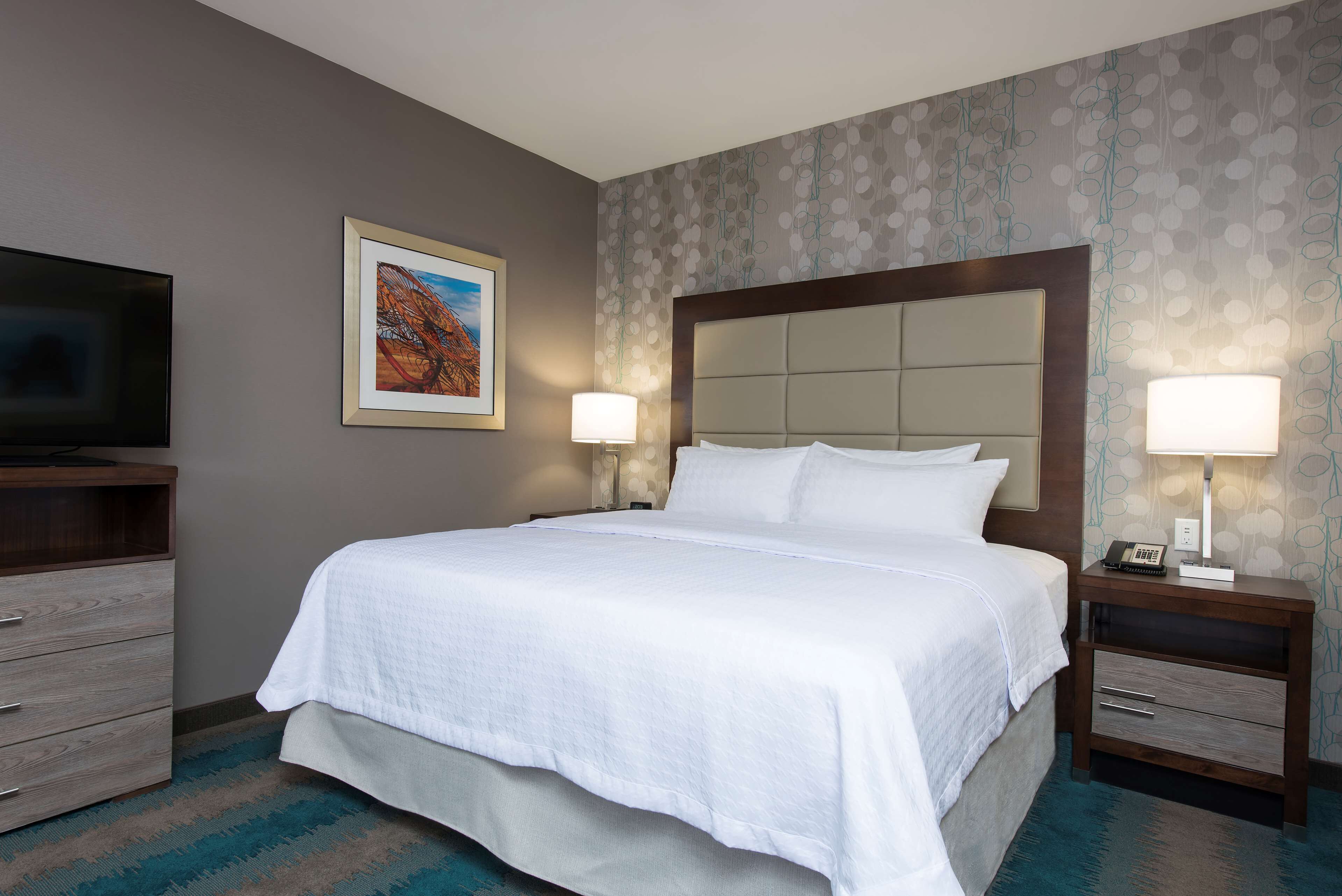 A neatly made bed with a headboard, a bedside table with a lamp, a TV on a stand, and a wall-mounted painting.