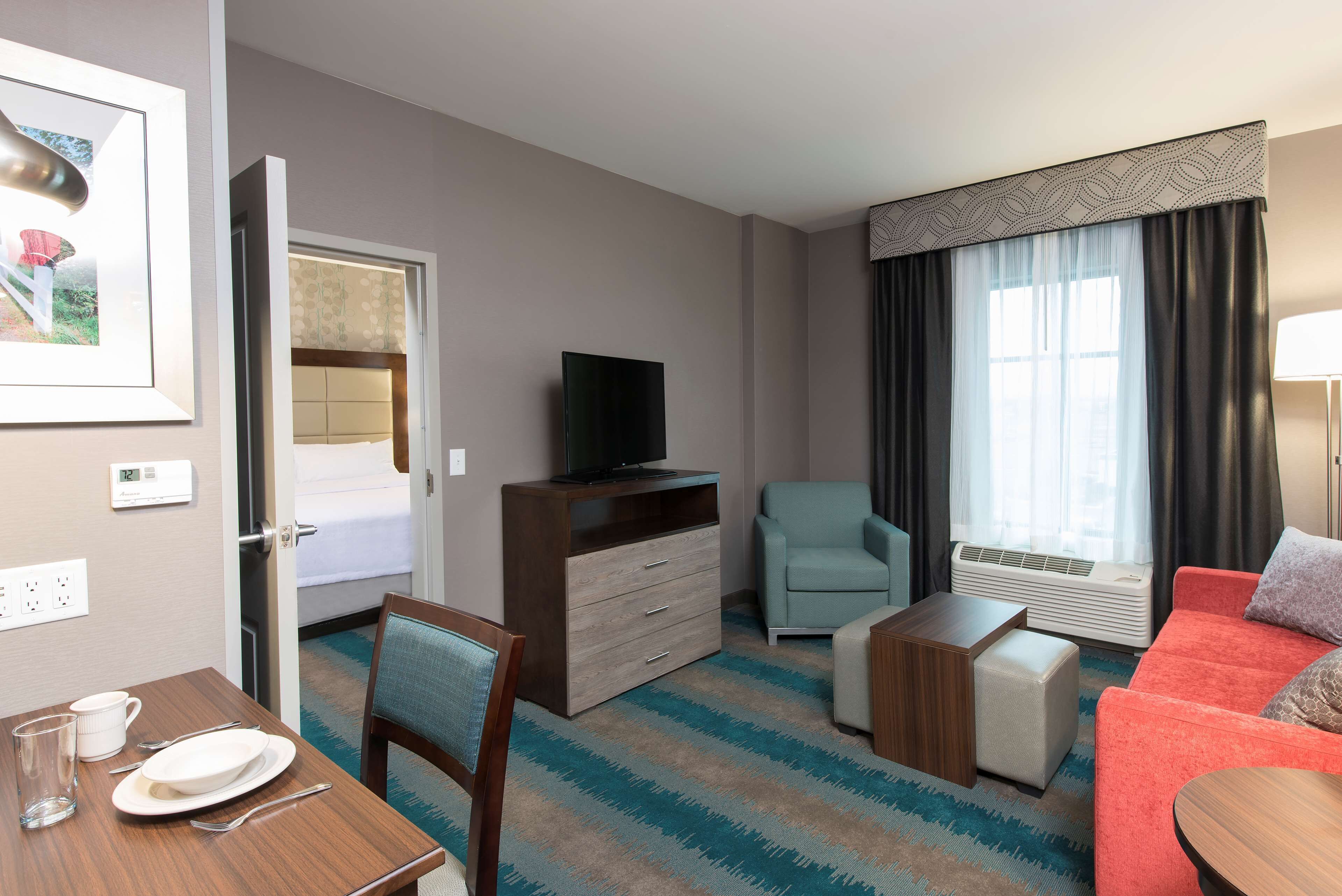 Una habitación de hotel con cama, televisión, sofá, sillón, mesa con plato y vaso, y ventana con cortinas.