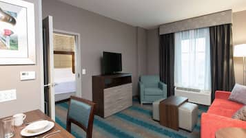 Quarto de hotel com cama, televisão, sofá, cadeira, mesa com prato e copo, e janela com cortinas.
