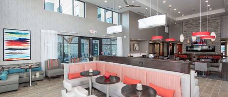 A spacious hotel lobby with a high ceiling, multiple seating areas, a bar, and a large TV screen.