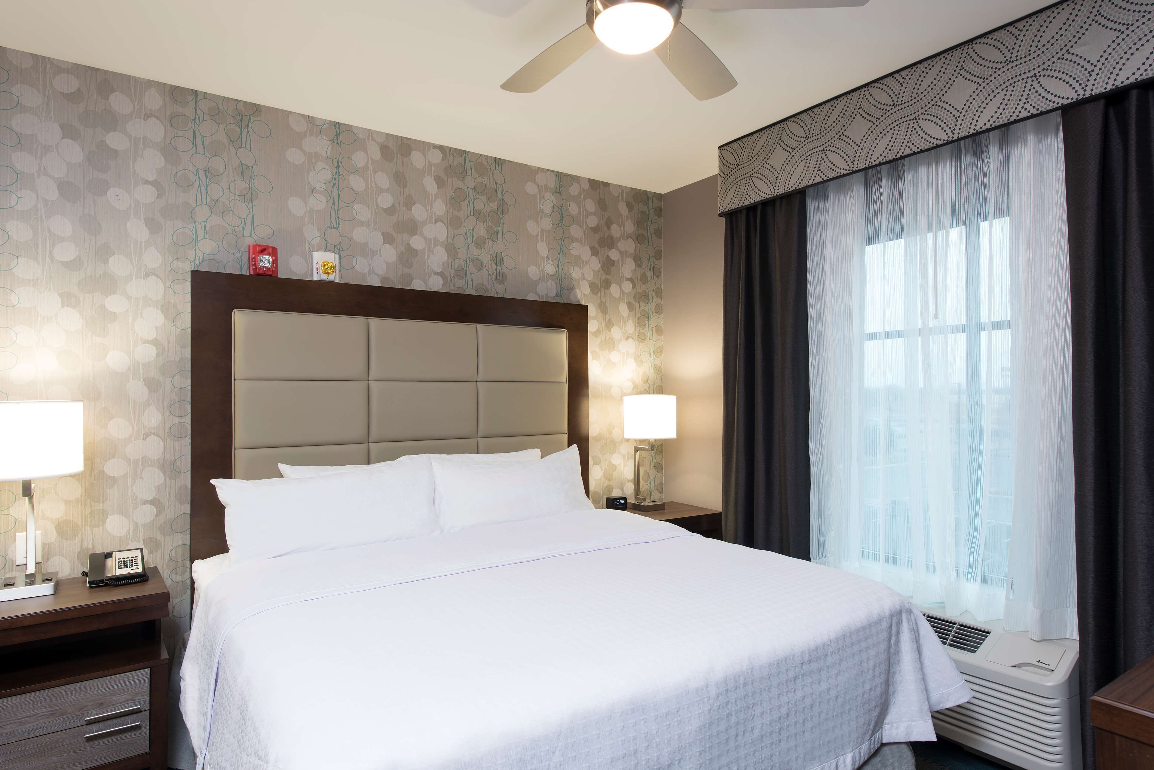 Une chambre d’hôtel avec un grand lit, deux tables de chevet avec des lampes, un ventilateur de plafond et une fenêtre avec des rideaux.