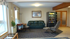 A living room with a green sofa, a grey armchair, a small table with a plant, a bookshelf, and a patterned rug. - Perfect Inns & Suites (Weyburn)