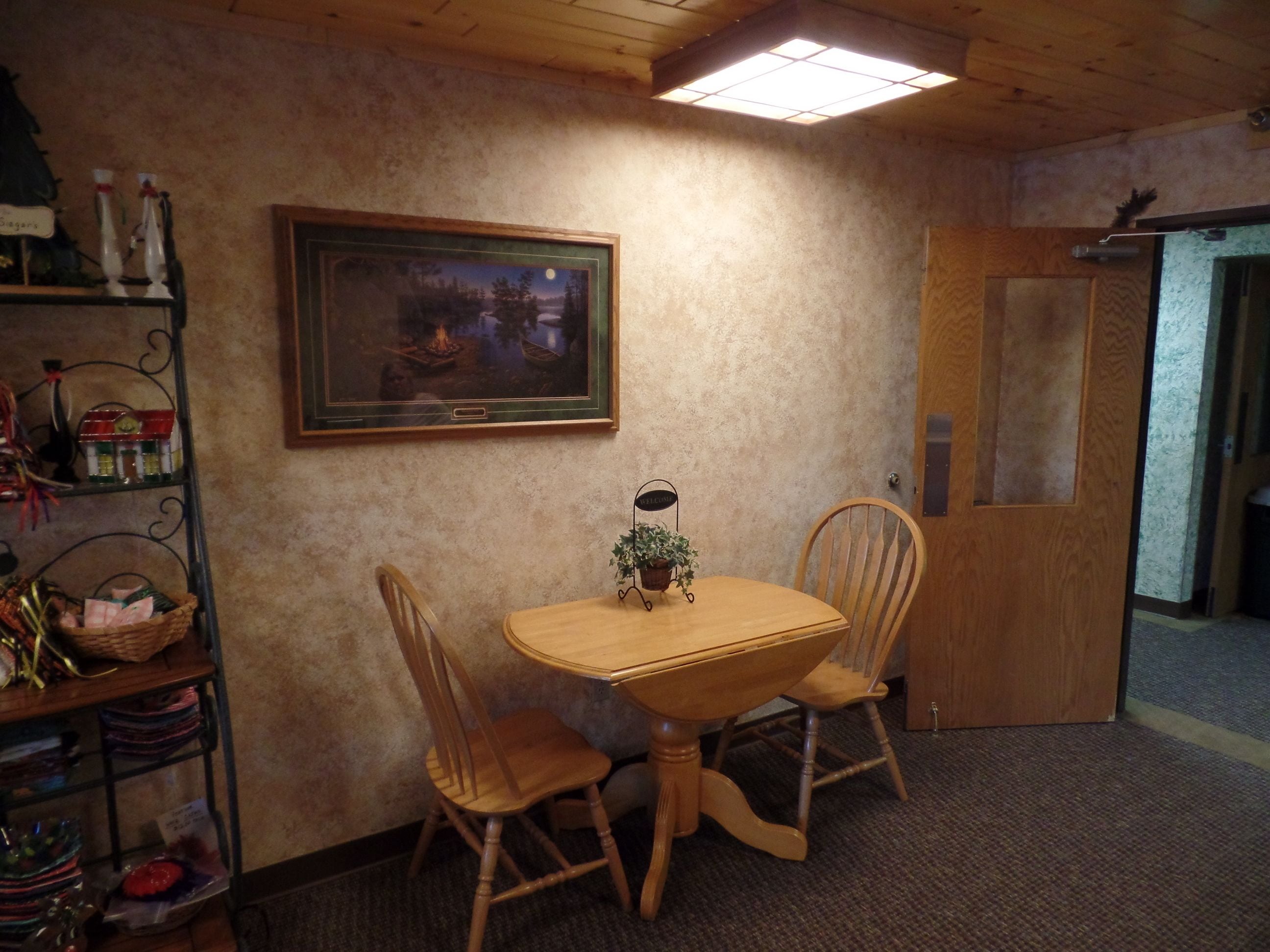 A room with a wooden dining table, two chairs, a painting of a night scene, a welcome sign, and a shelf with various items.