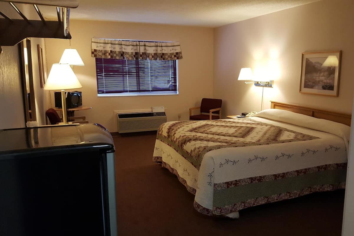 A hotel room with a bed, two bedside lamps, a chair, a heater, a window with blinds, and a painting on the wall.
