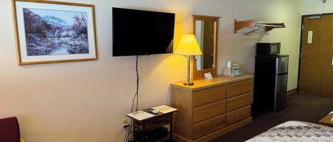 A hotel room with a bed, a TV mounted on the wall, a wooden dresser, a mirror, a lamp, and a refrigerator.