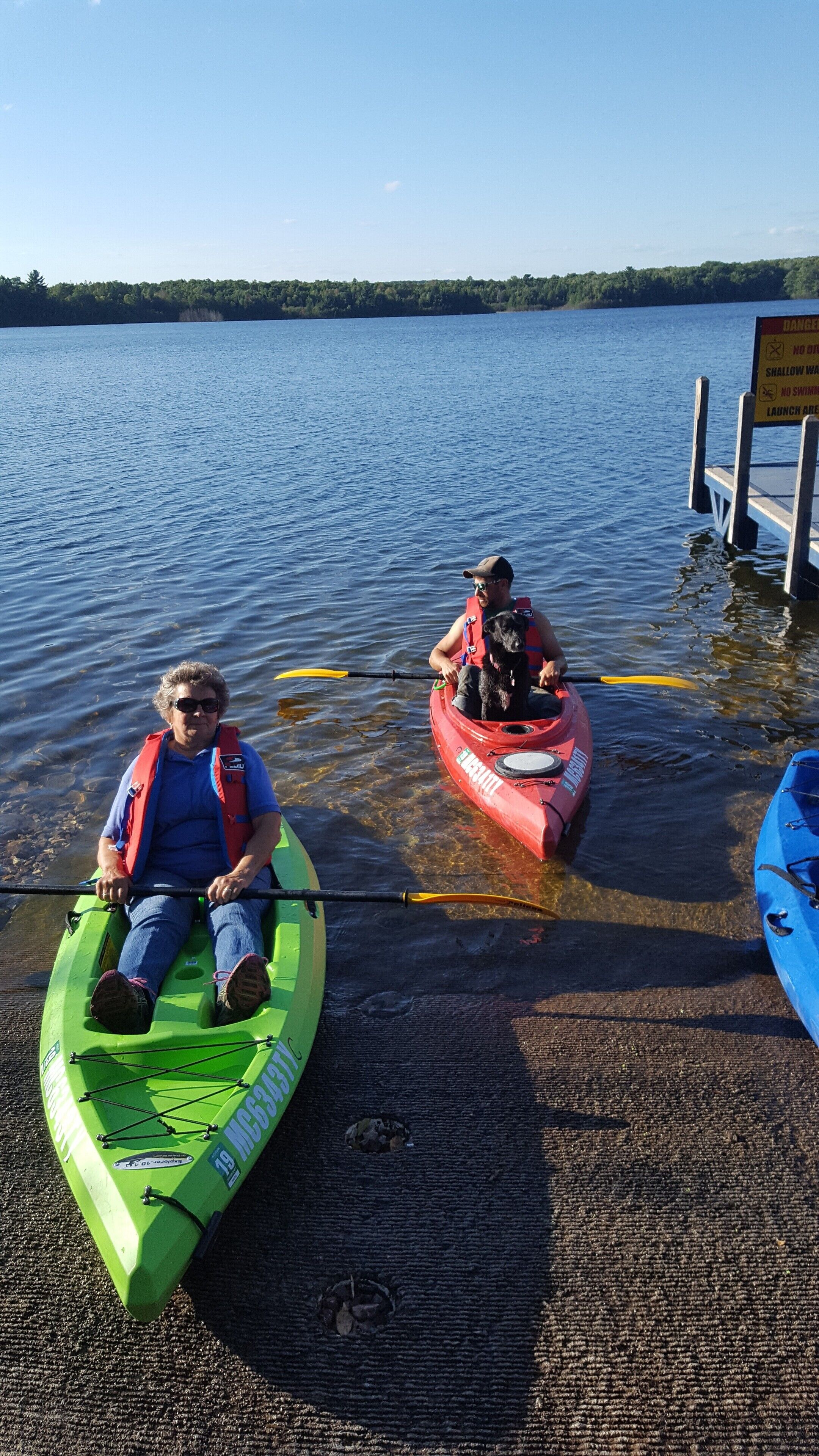 Two people in kayaks at a dock with a sign warning about shallow water and no swimming.