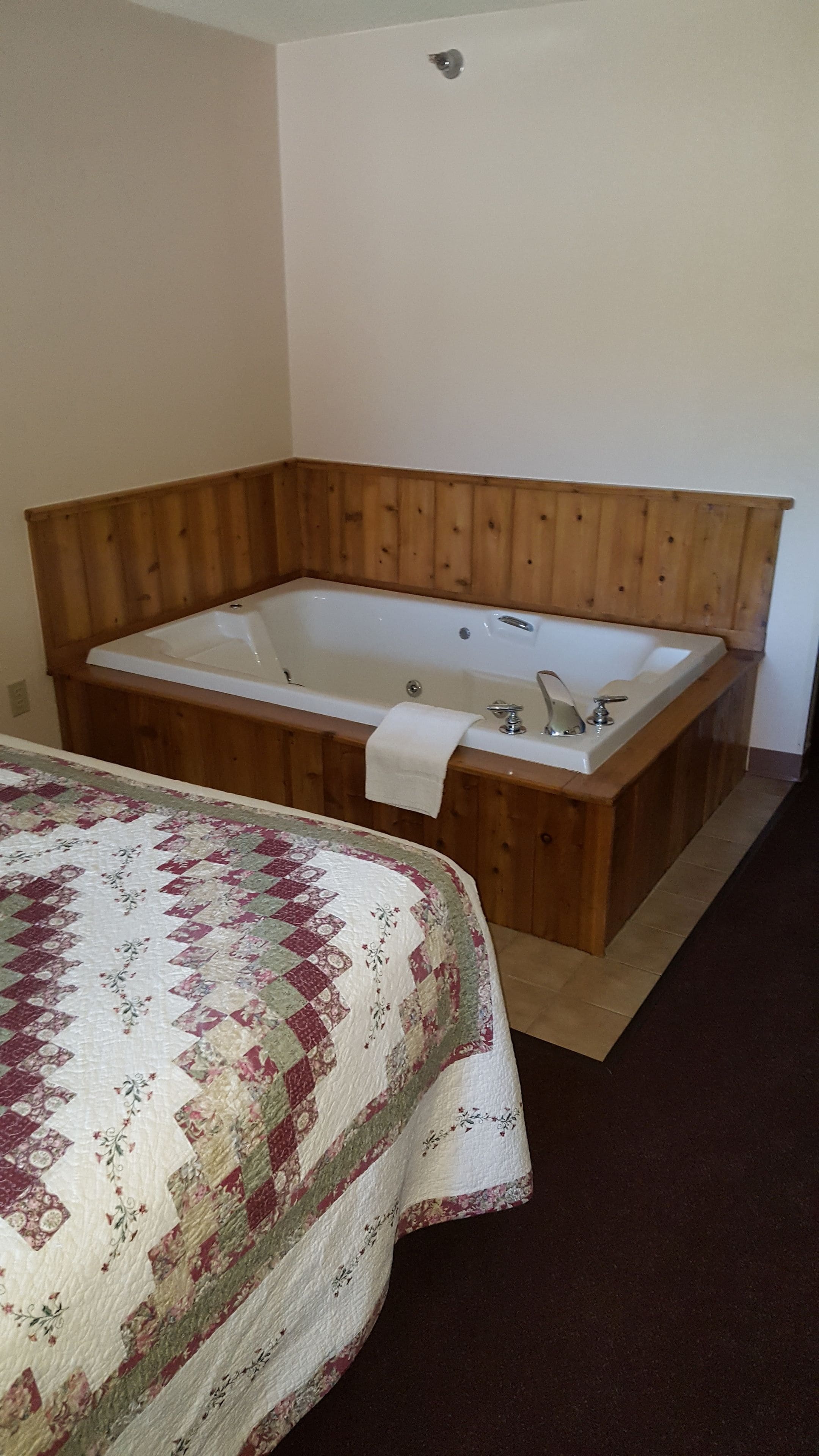 A bathroom with a wooden bathtub, a quilted bedspread, and a wooden floor.