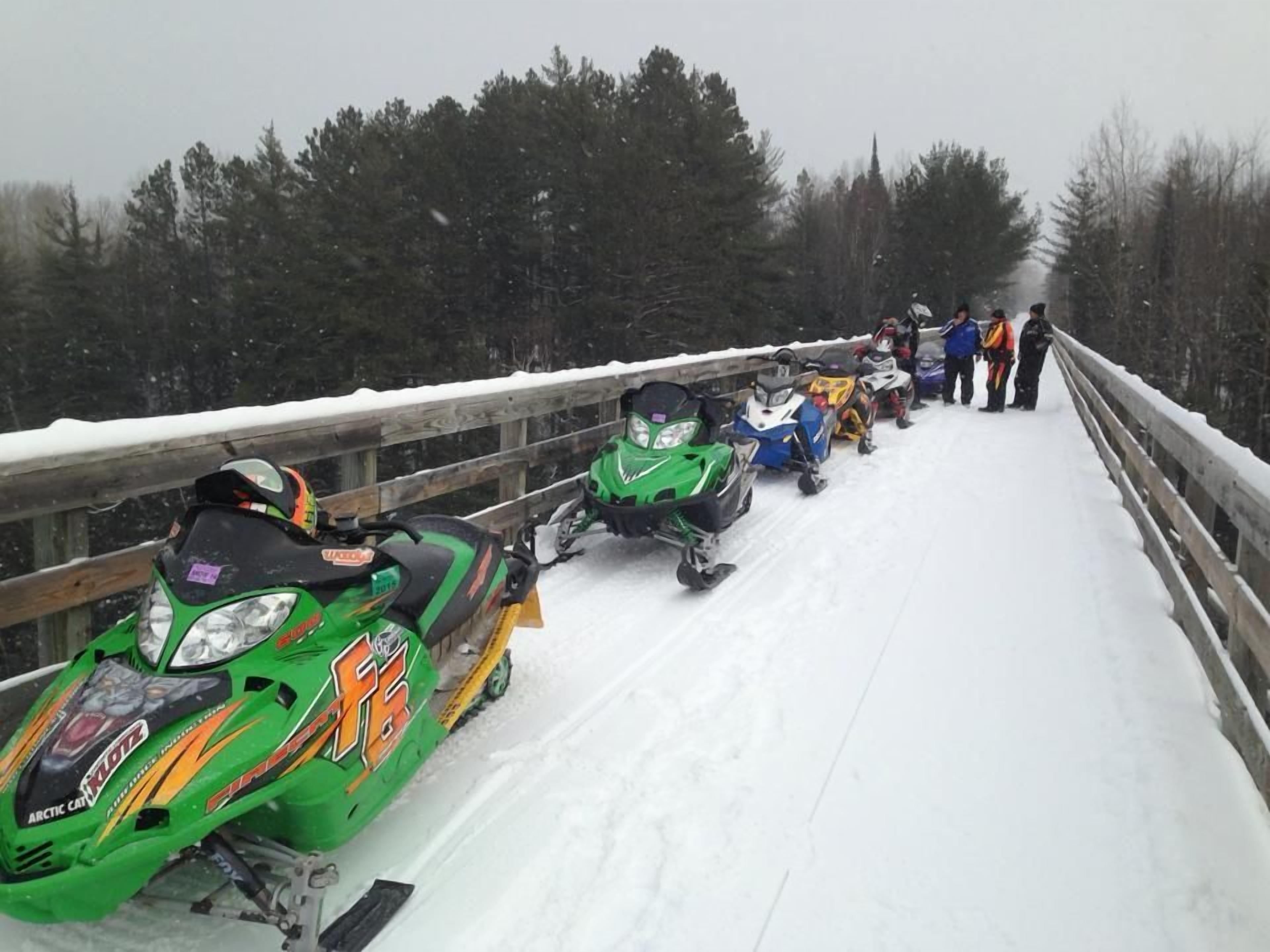 A line of snowmobiles parked on a snowy bridge.