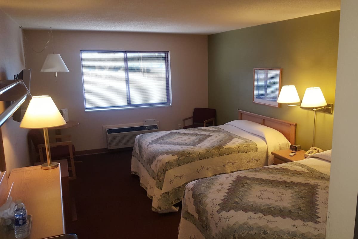 A hotel room with two beds, a desk, a chair, a lamp, and a window with blinds.