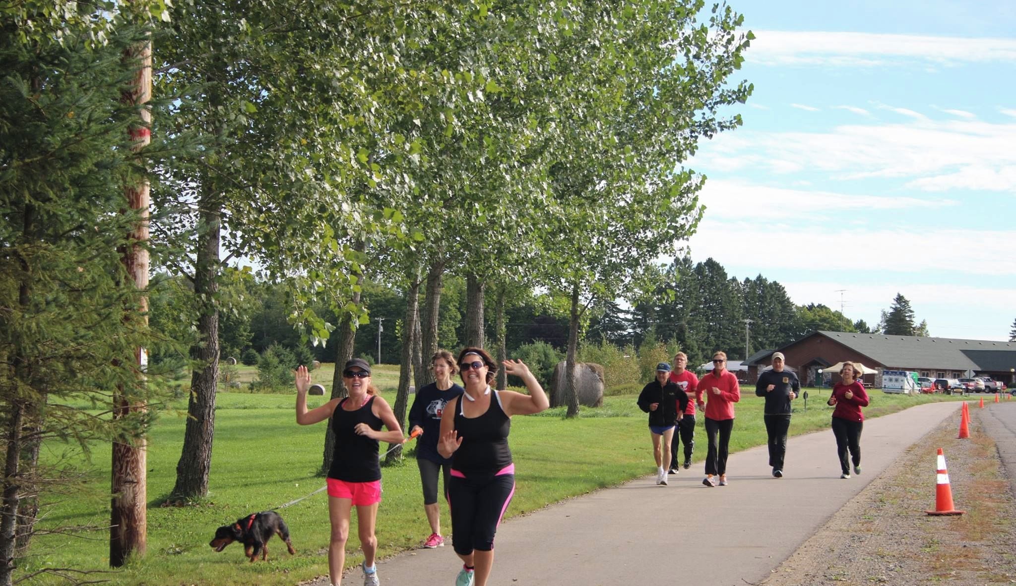 Eine Gruppe von Menschen joggt auf einem gepflasterten Weg in einem Park mit Bäumen und einem Gebäude im Hintergrund.