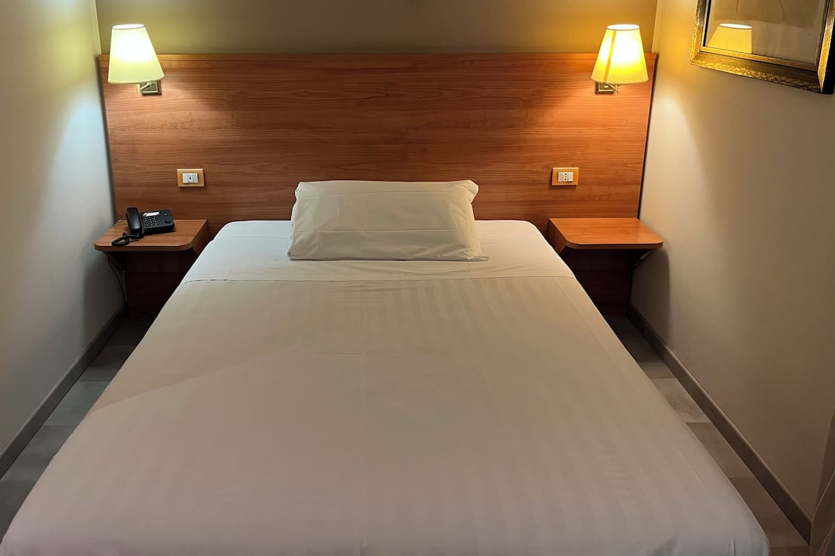 Une chambre d’hôtel avec une tête de lit en bois, deux lampes de chevet, un téléphone et un lit bien fait, recouvert de draps blancs.