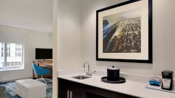 A hotel room with a white sink, a framed cityscape artwork, a coffee maker, and a blue chair.