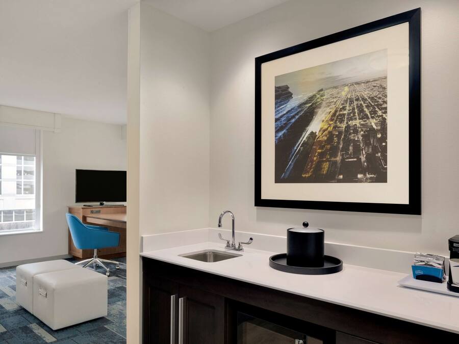 A hotel room with a white sink, a framed cityscape artwork, a coffee maker, and a blue chair.