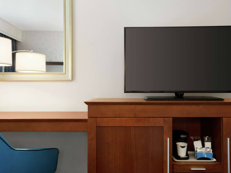 Quarto de hotel com televisão de tela plana presa a um armário de madeira, uma cadeira azul e uma cafeteira.