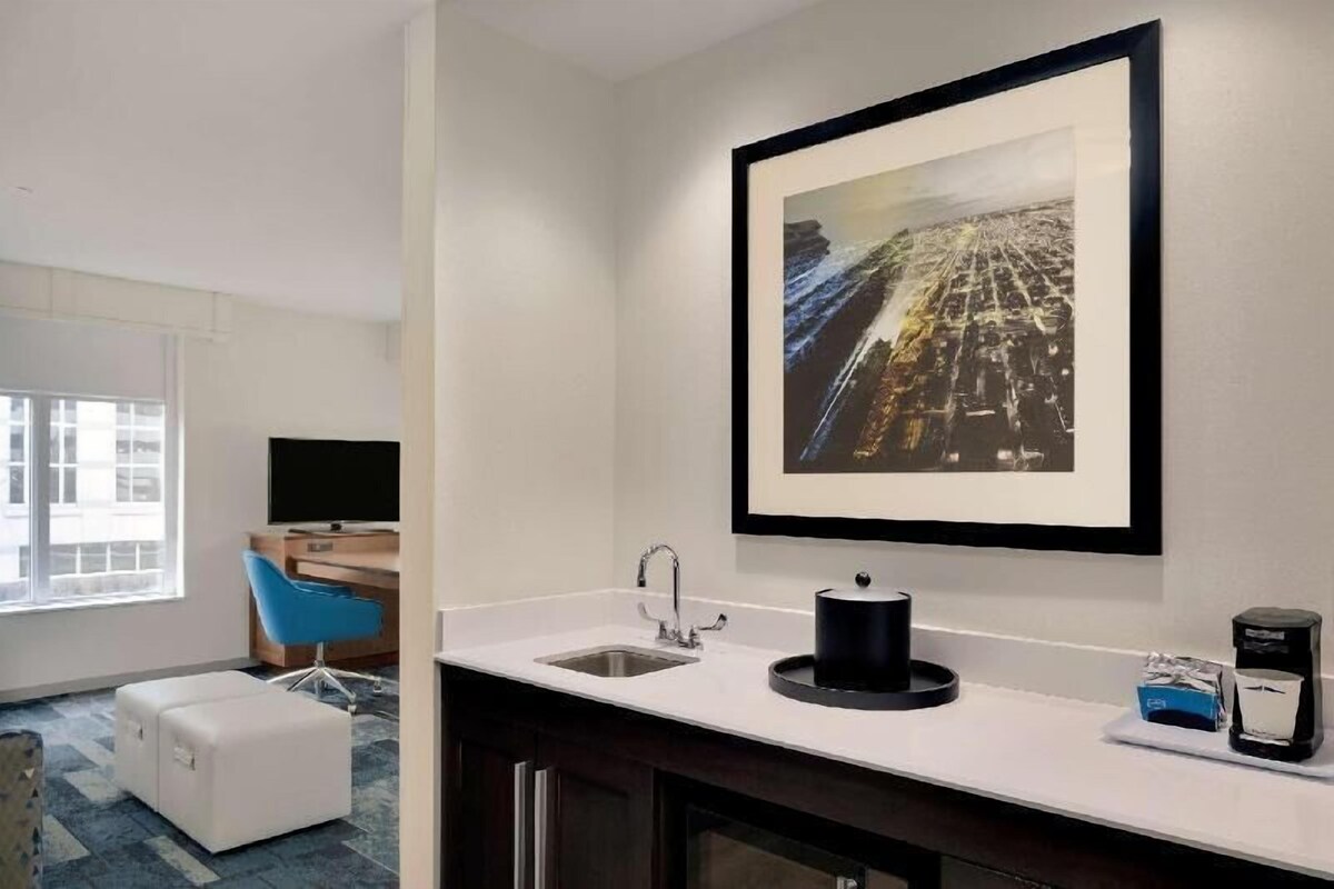 A modern hotel room with a white sink, a framed cityscape artwork, a blue chair, and a desk with a flat-screen TV.