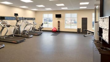 Een sportschool met loopbanden, een hometrainer, een roeimachine, een rode fitnessbal en een aan de muur bevestigde televisie.