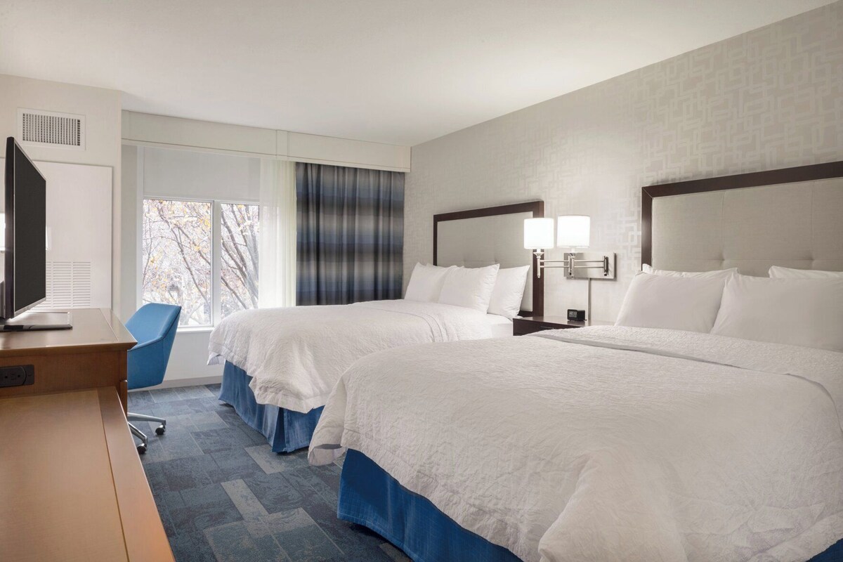 A hotel room with two beds, a desk, a chair, a TV, and a window with curtains.