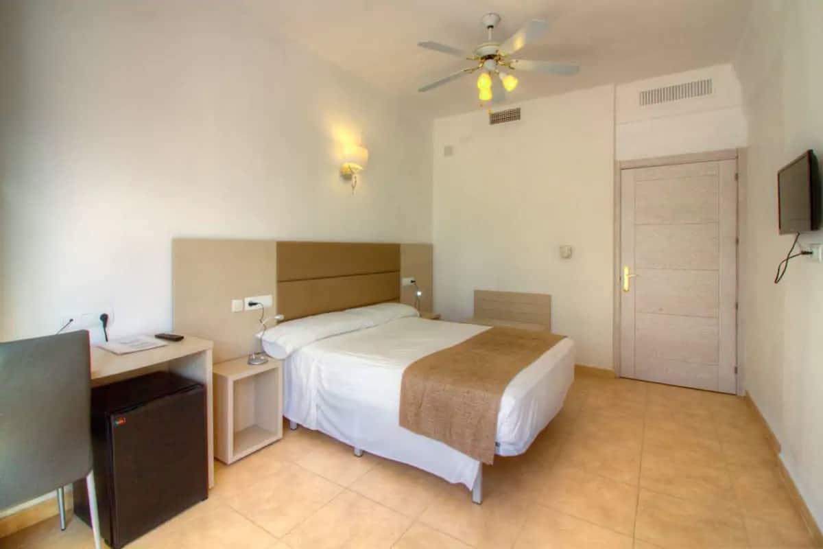 Un dormitorio con cama, escritorio, silla, ventilador de techo, televisor y puerta.