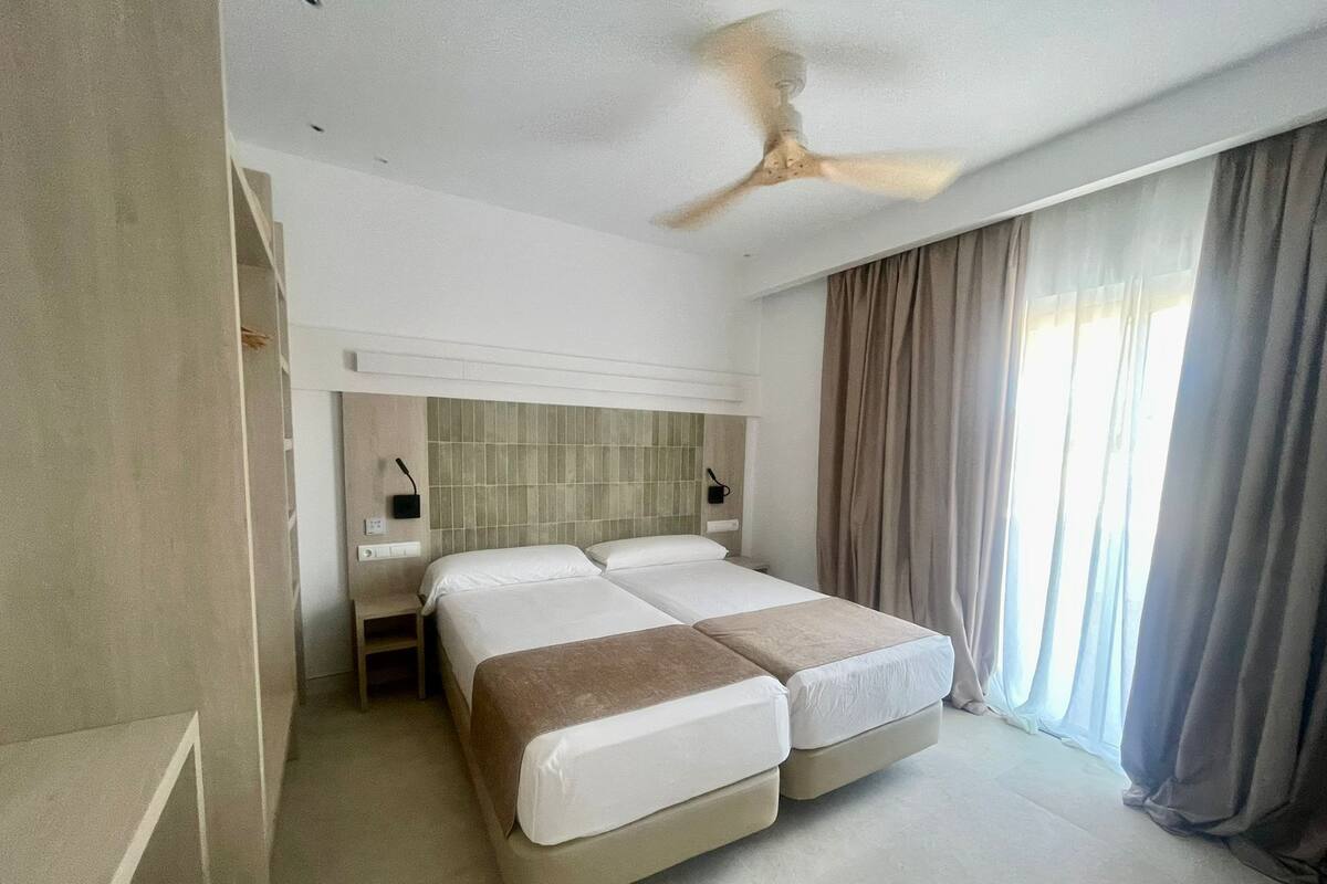 Habitación de hotel con una cama grande, un ventilador de techo, una mesita de noche y una ventana con cortinas.