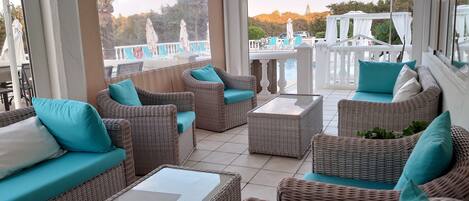 A sunlit outdoor seating area with wicker furniture and a glass-top table, overlooking a pool and a white fence.