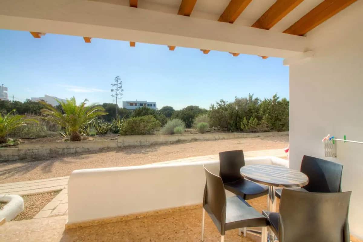 Un espacio al aire libre cubierto con una mesa y sillas, con vistas a un jardín ajardinado y edificios a la distancia.